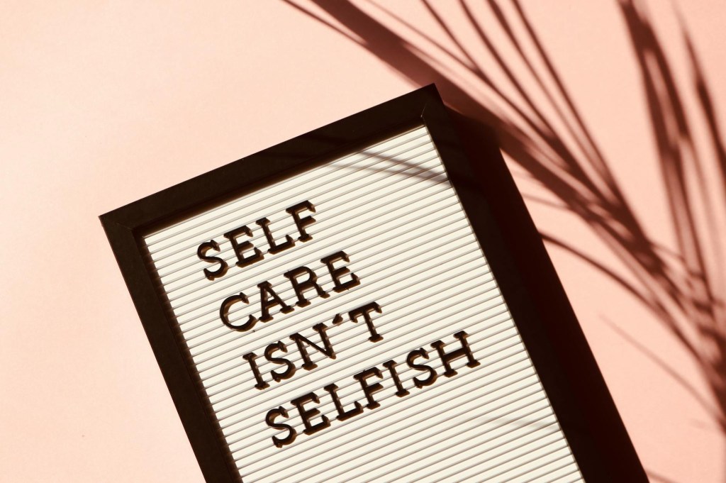 A framed sign with the text 'SELF CARE ISN'T SELFISH' displayed against a pink background, accompanied by a shadow from a plant.